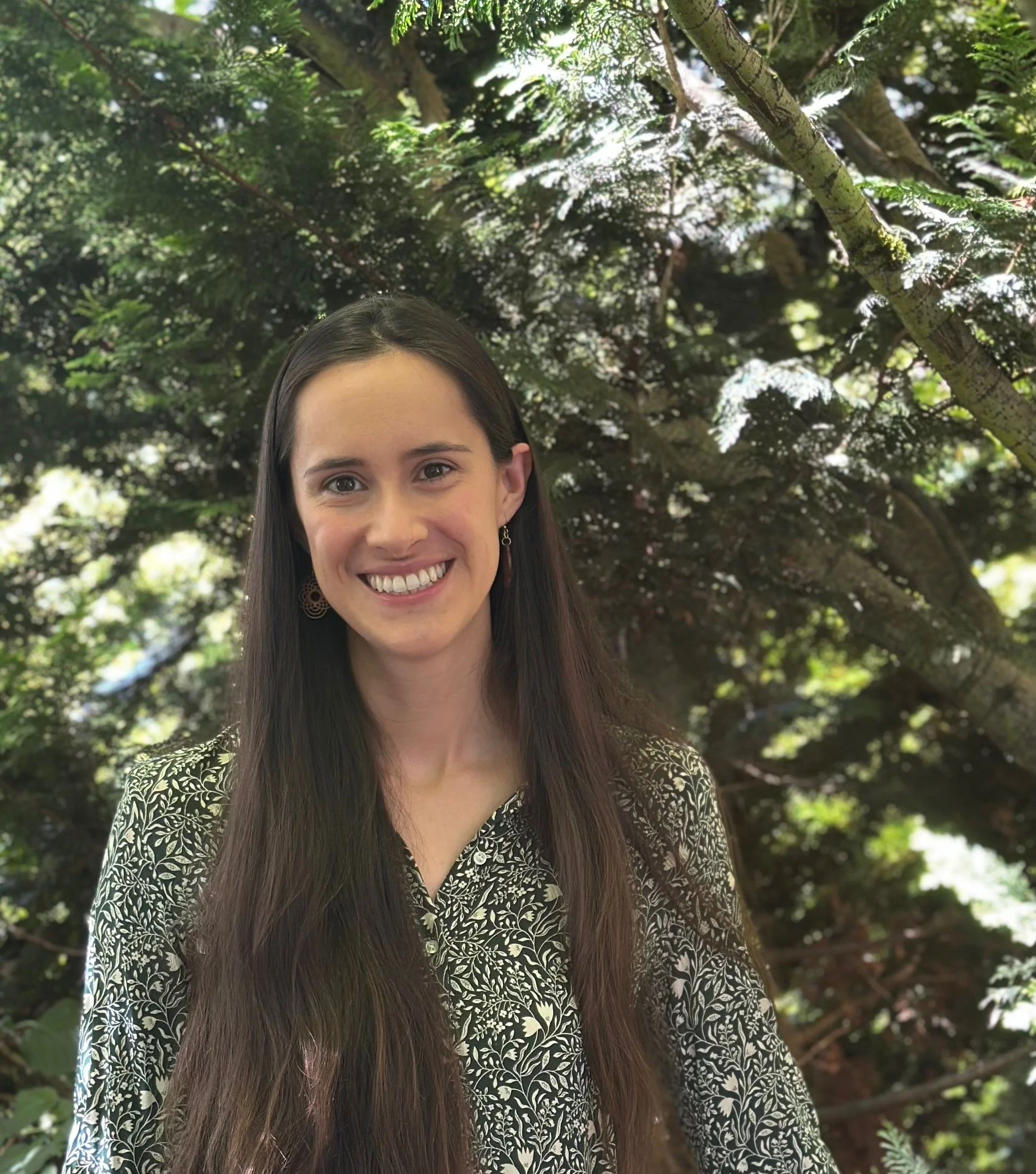 A woman with long dark hair smiling outdoors in front of green trees with sunlight through the leaves.