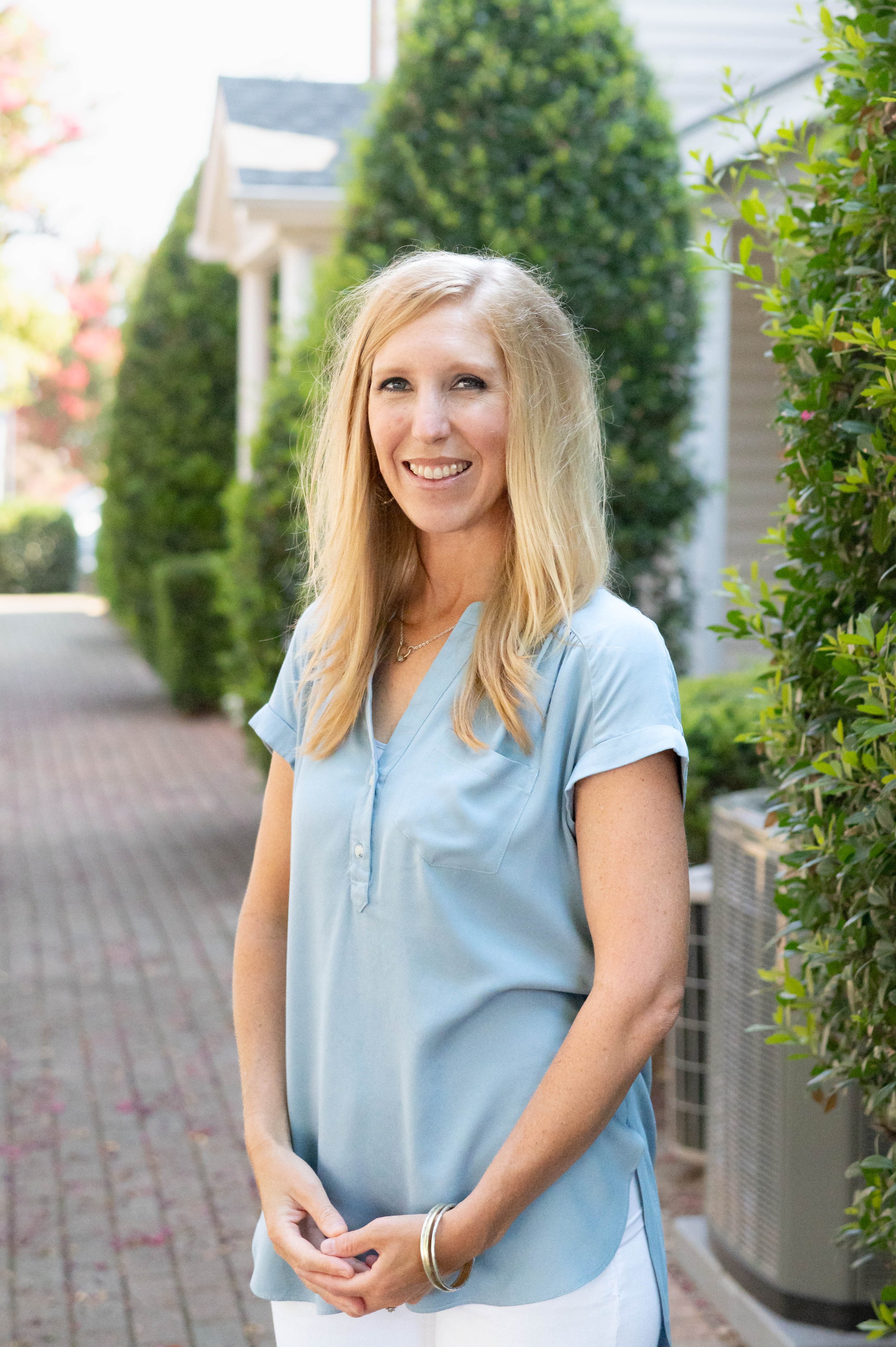A woman with long blonde hair and a light blue blouse standing outdoors on a brick sidewalk with green bushes and trees in the background.