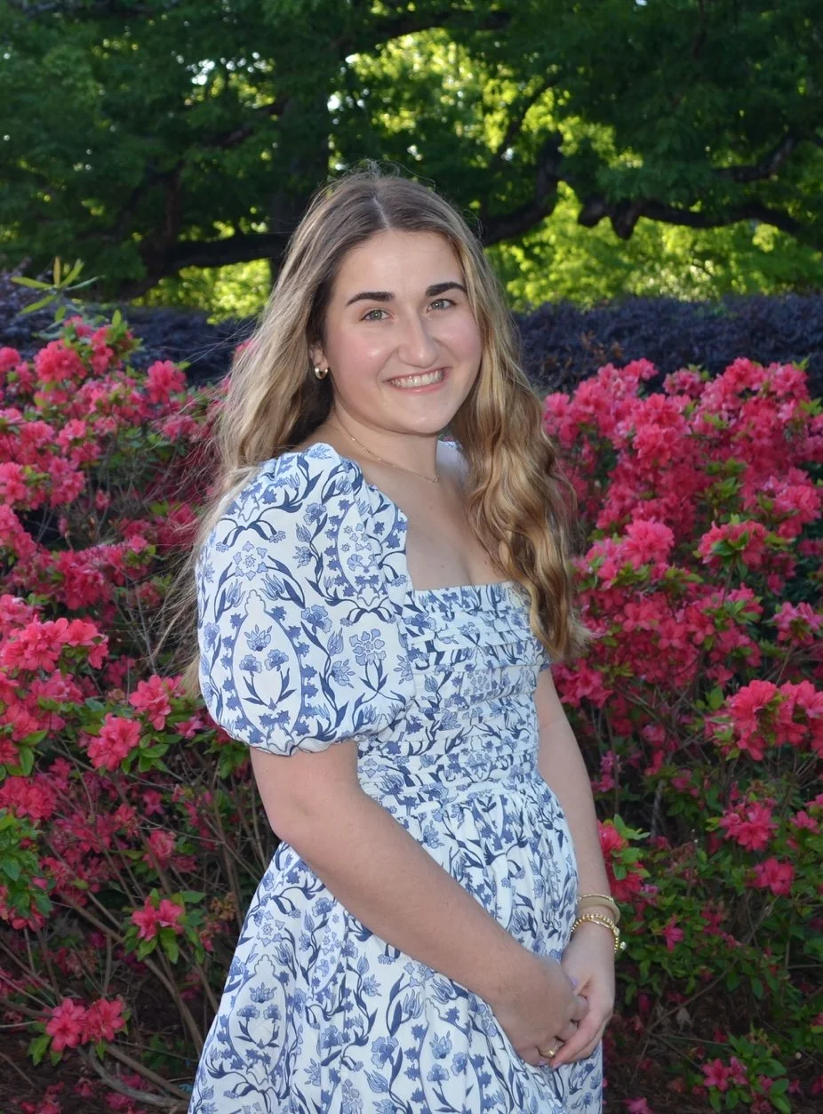 A young woman with long wavy hair wearing a blue and white floral dress, standing outdoors in front of pink flowering bushes and green trees.