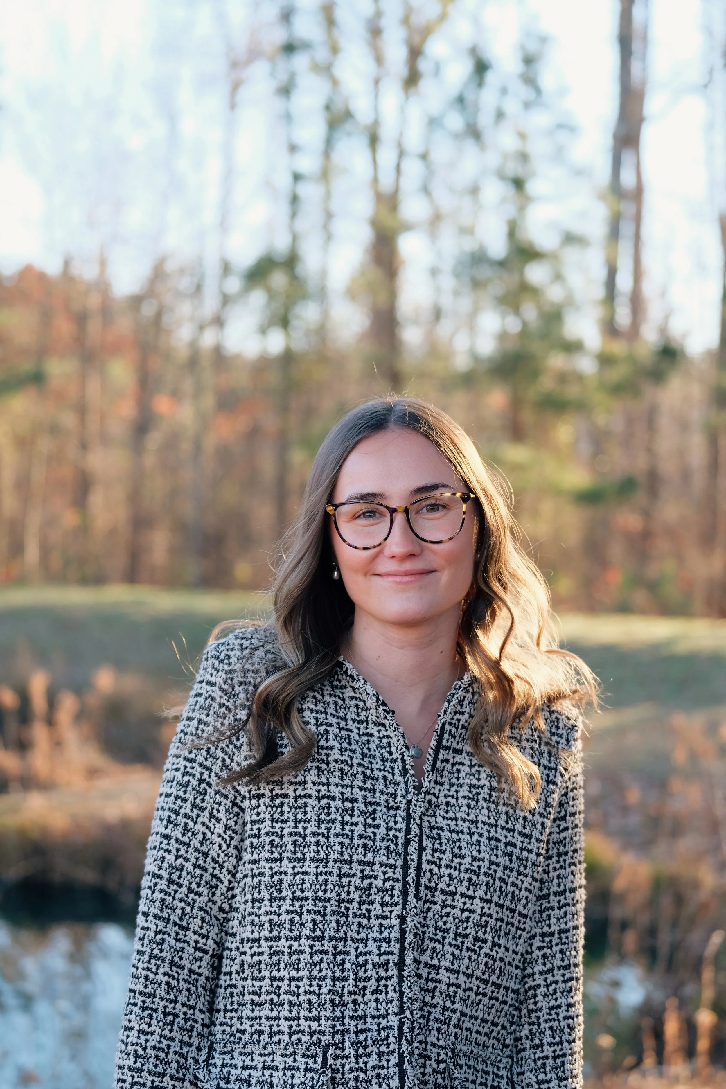 A young woman with glasses and wavy hair standing outdoors in front of a background of trees and a pond, smiling softly at the camera.