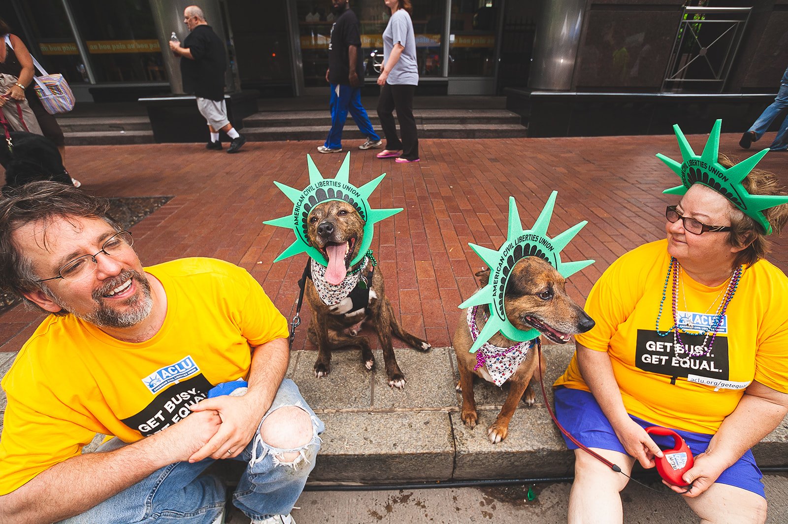 Man sits on Liberty Avenue with mother-in-law in yellow "Get Busy, Get Equal" shirts from the ACLU during Pittsburgh Pride. Two jubilant dogs sit between them wearing ACLU liberty crowns.