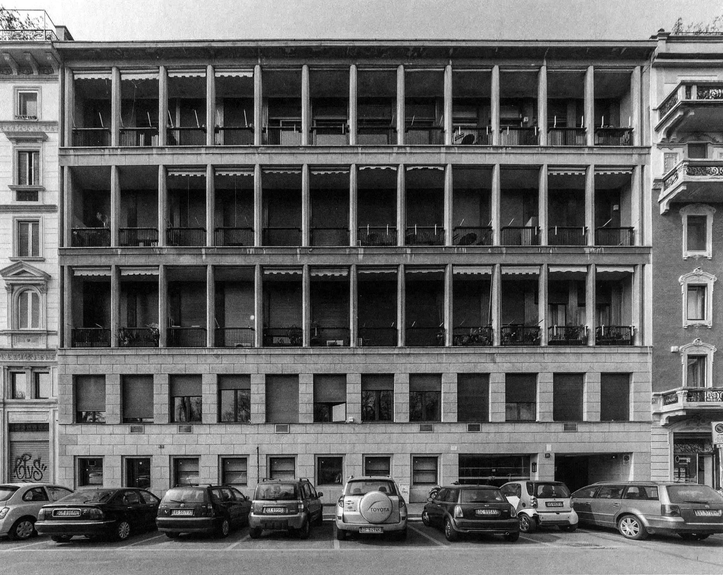 A black and white photograph of a multi-story building with a modern architectural design, featuring open balconies with vertical columns, and several cars parked in front of it on the street.