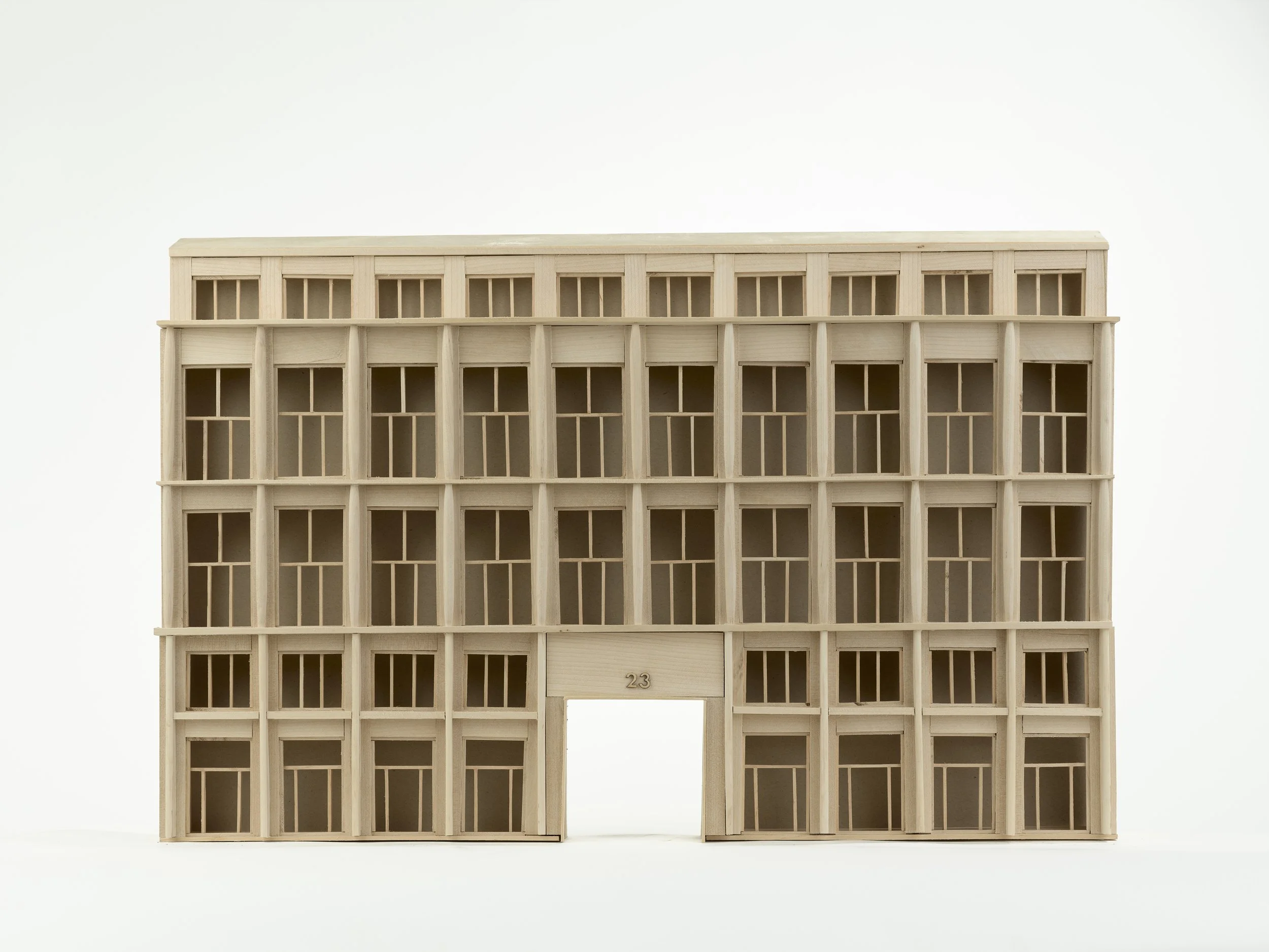 A wooden model of a building with several rectangular windows and a door, and the number 23 above the entrance, set against a plain white background.