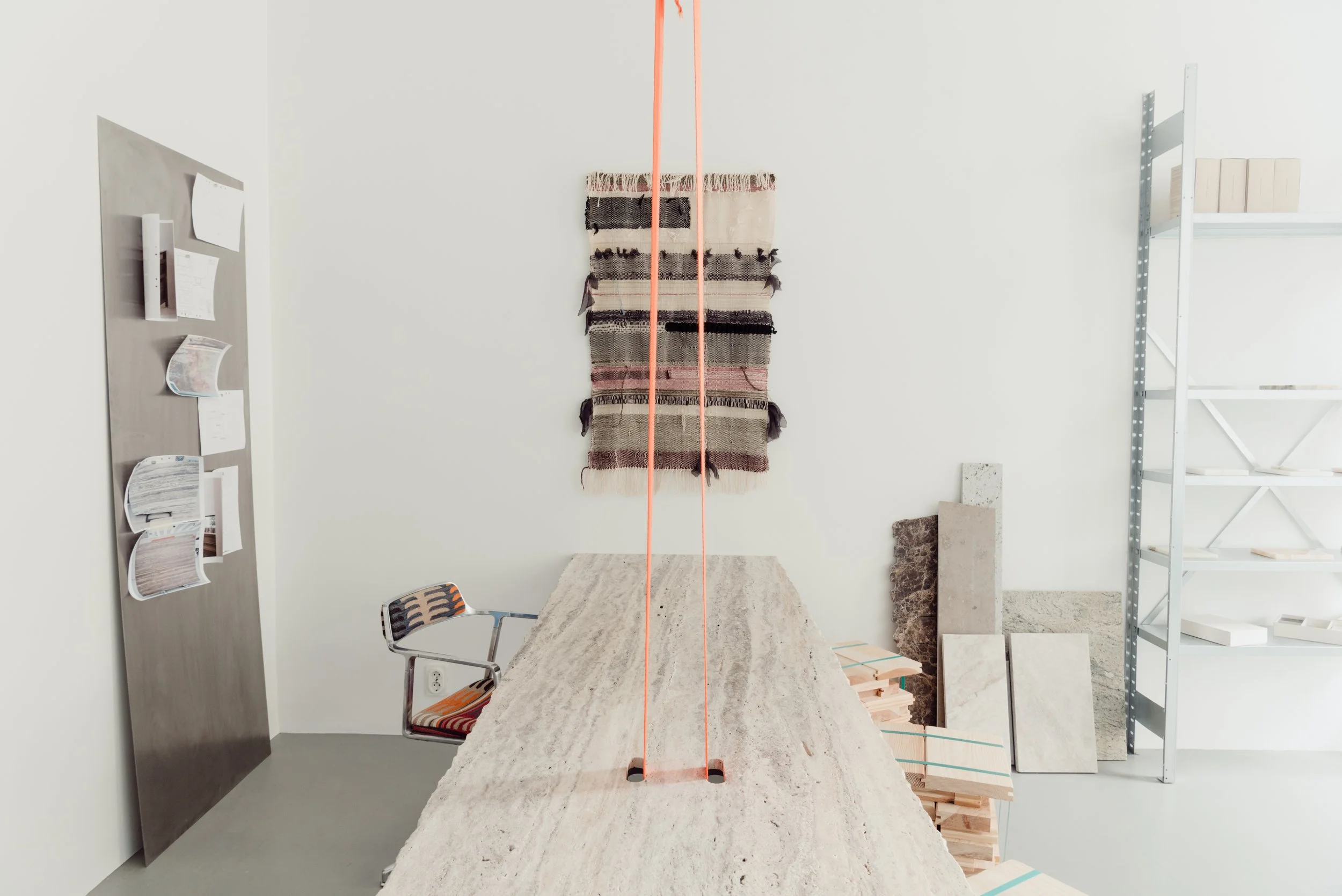 Interior of a space with a large rectangular stone or concrete table, two chairs, a wall with hanging textiles and shelves with materials, and construction tools, suggesting a workspace or studio.
