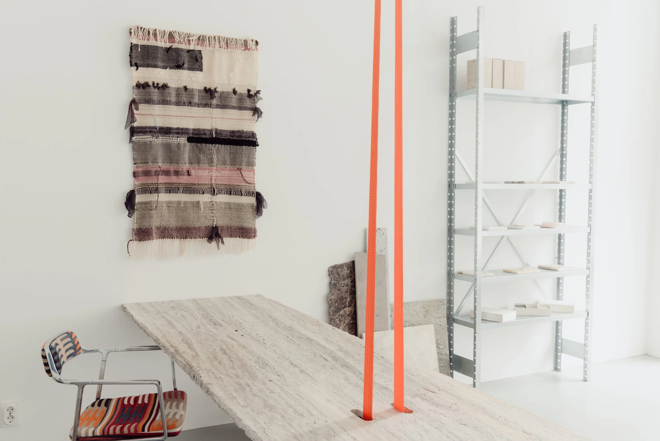 Interior of an art studio or workspace with a white wall, a textile art piece hanging on the left, a textured bench or table, two orange vertical poles in the middle, and a metal shelving unit on the right holding books and materials.