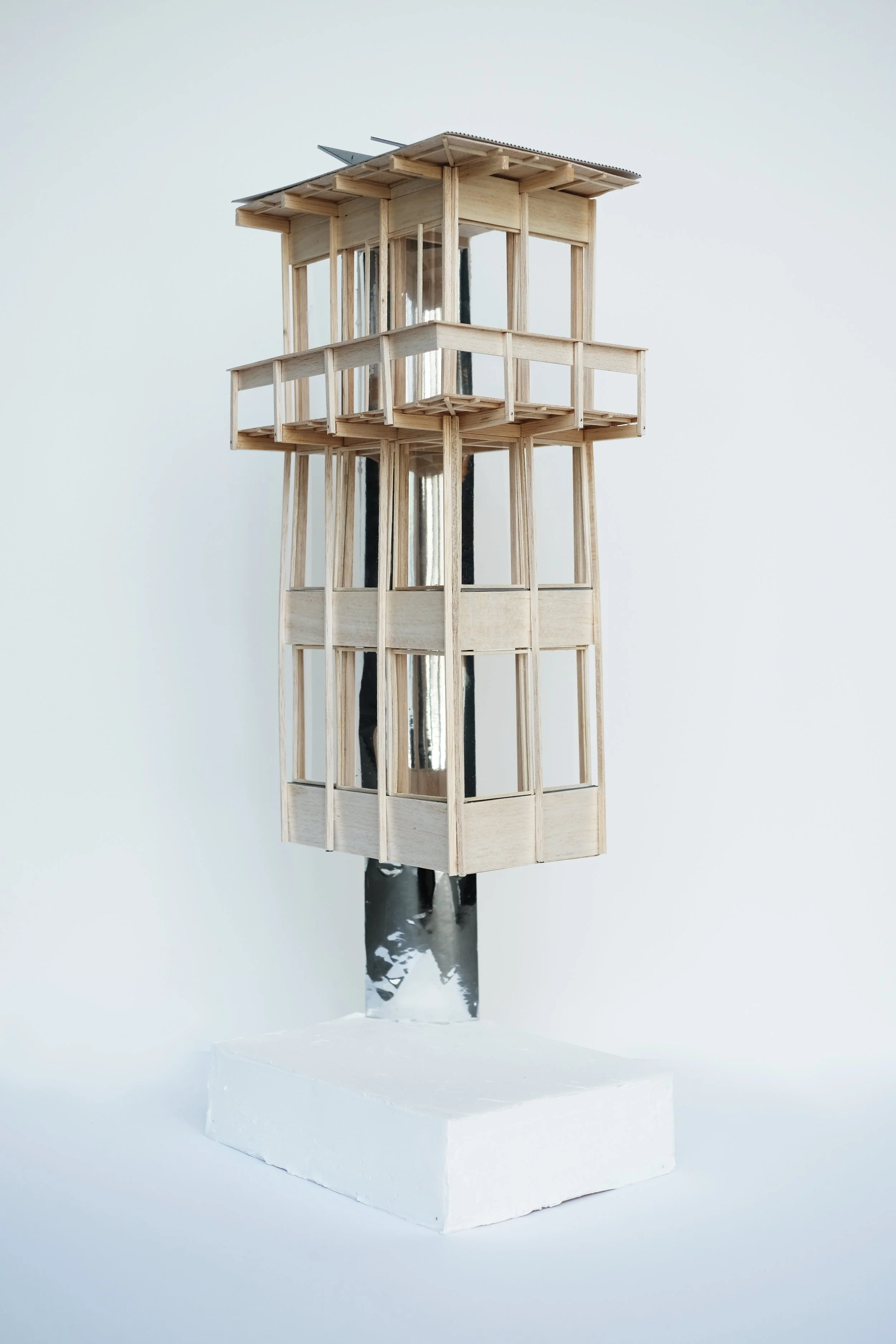 A small wooden model of a multi-level observation tower mounted on a square piece of white foam.