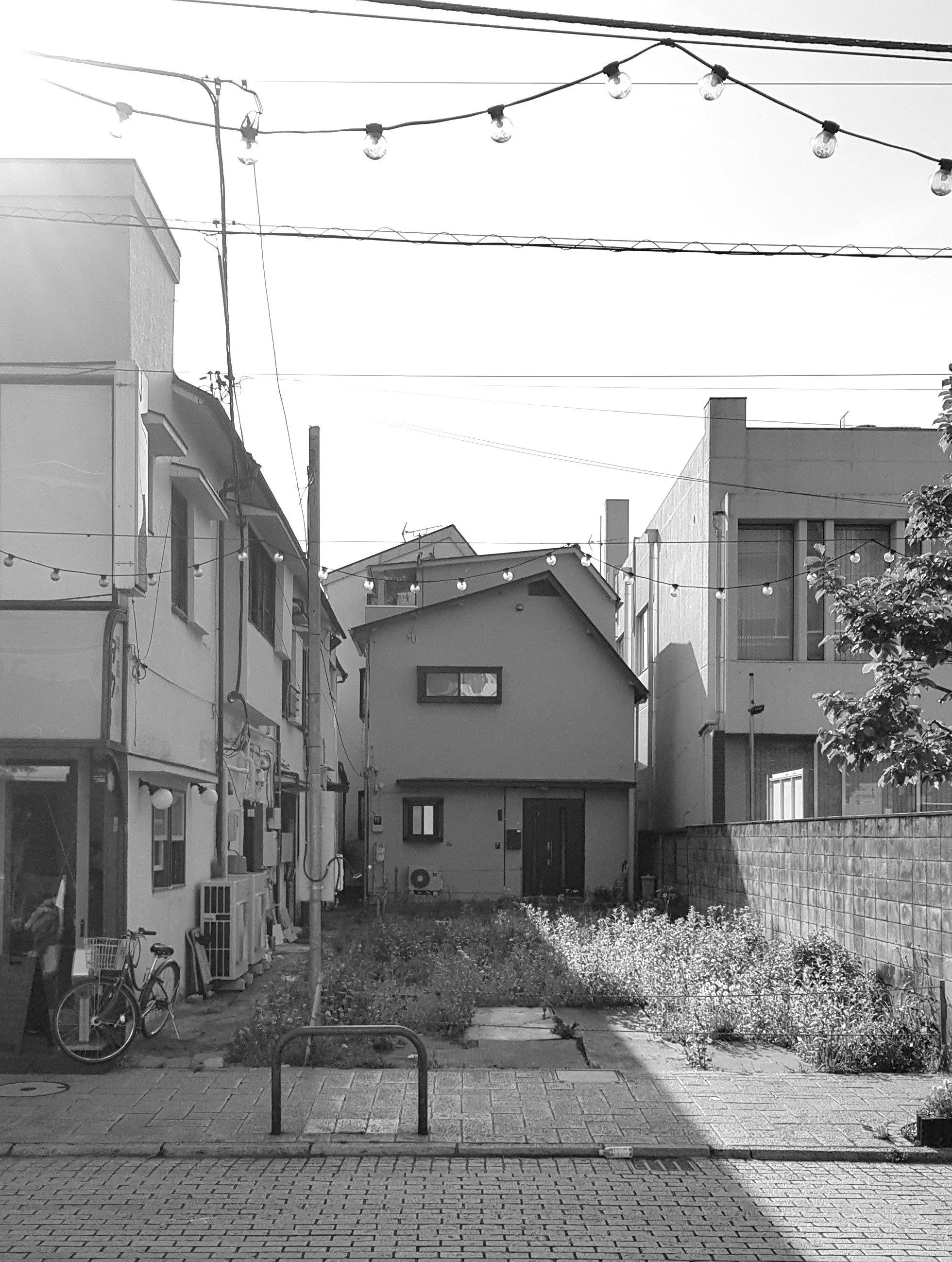 A black and white photograph of a small backyard surrounded by buildings. The yard has a patch of grass and a small garden bed, bordered by a brick pathway. Overhead, string lights are hanging between the buildings. On the left side, there is a bicyc