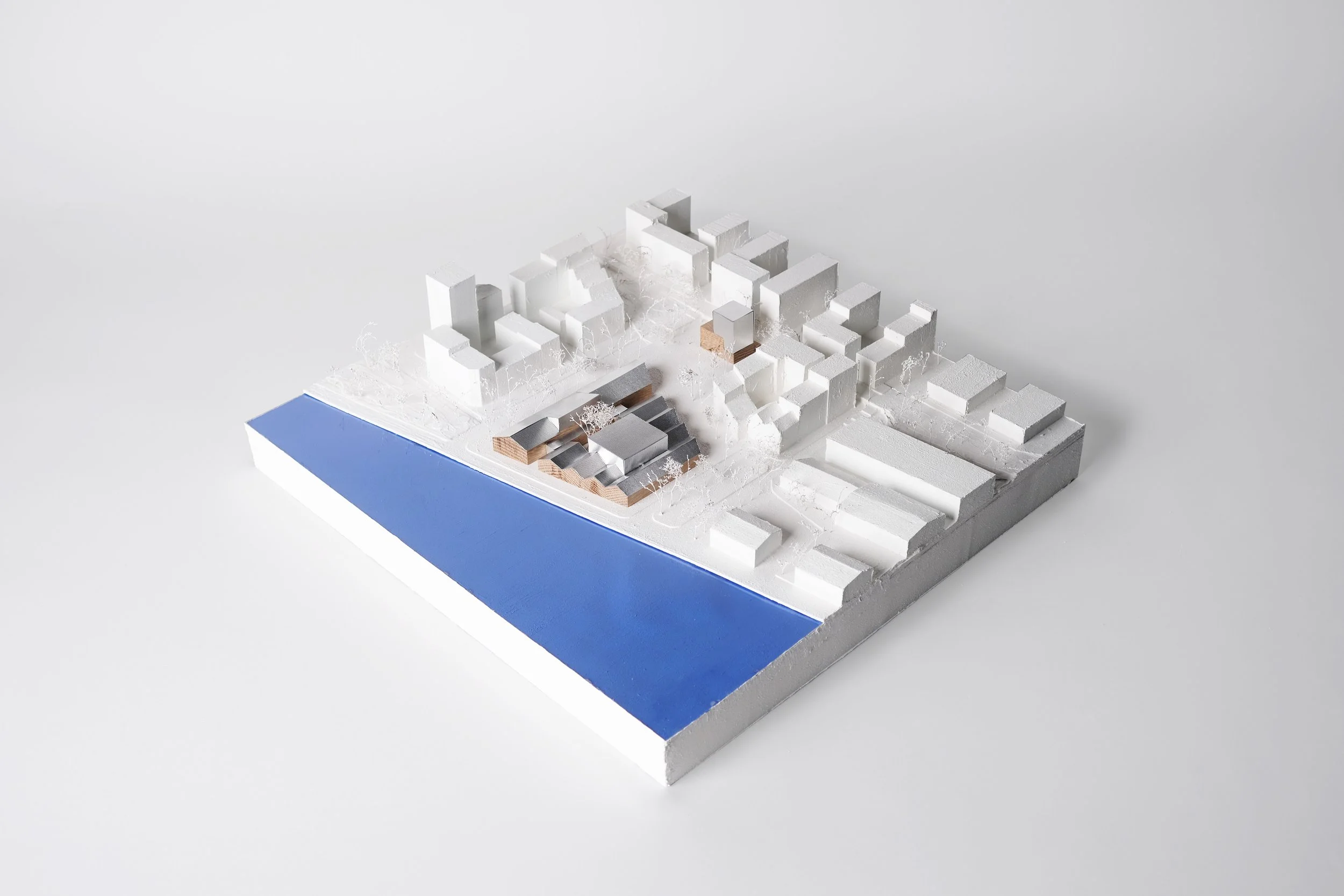 Architectural model of a building complex featuring white structures, some with flat roofs and others with pitched roofs, situated on a base with a blue section representing water and white pathways or roads. There are also miniature trees dispersed 