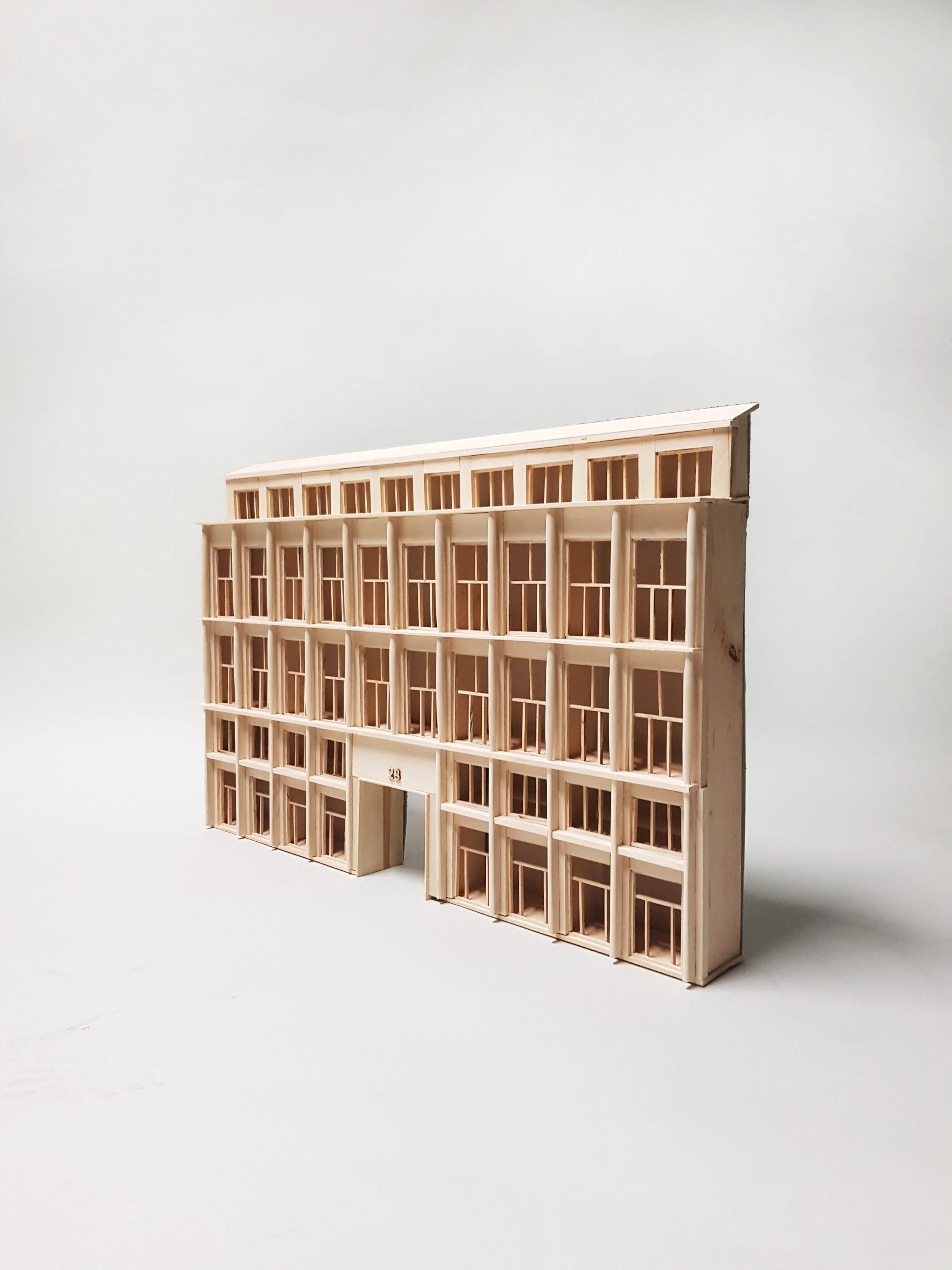 A miniature wooden model of a multi-story building with detailed window frames and a small entrance, set against a plain white background.