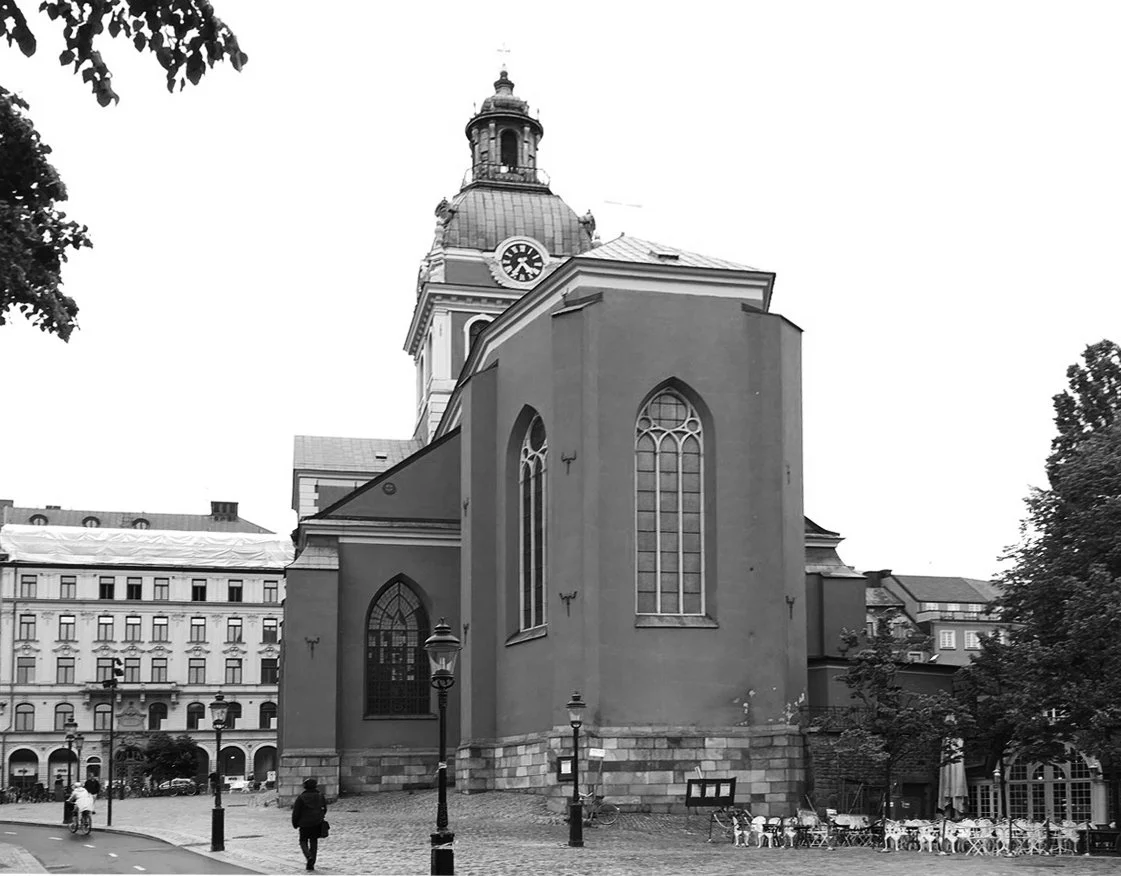 A black-and-white photo of a historic church with a tall clock tower and large arched stained glass windows, located in a city square with street lamps, benches, and a few pedestrians.