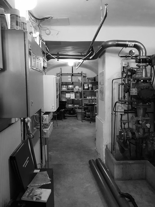 A black and white photo of a basement or utility room with pipes, shelving with various boxes and items, and machinery.