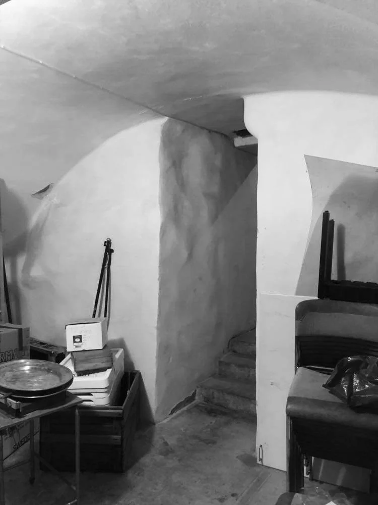 Interior view of a basement or cellar with curved walls and stairs leading upward, cluttered with storage boxes, a scale, stacked chairs, and miscellaneous items.