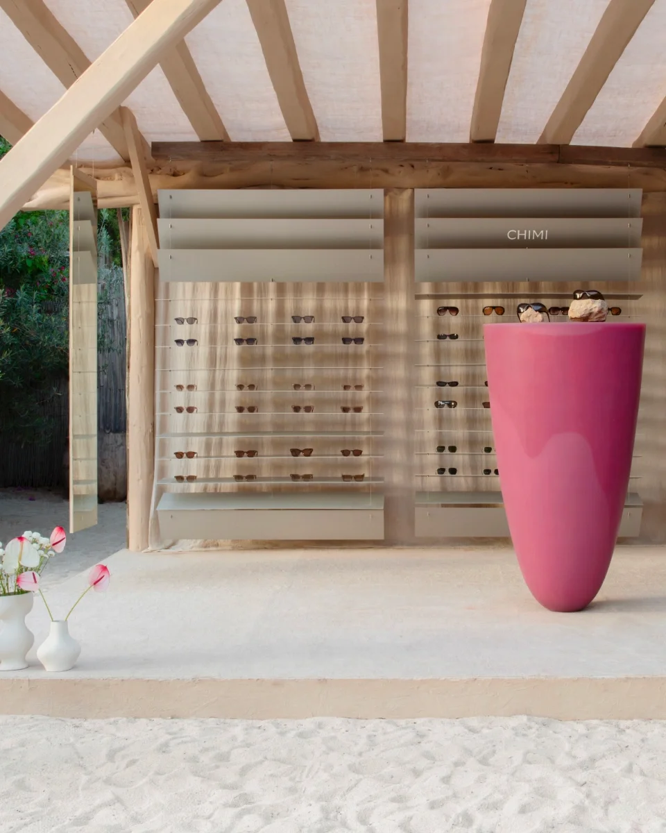 Display of sunglasses on a wooden slat wall with a pink flowerpot in front, located inside a beachside or outdoor structure.