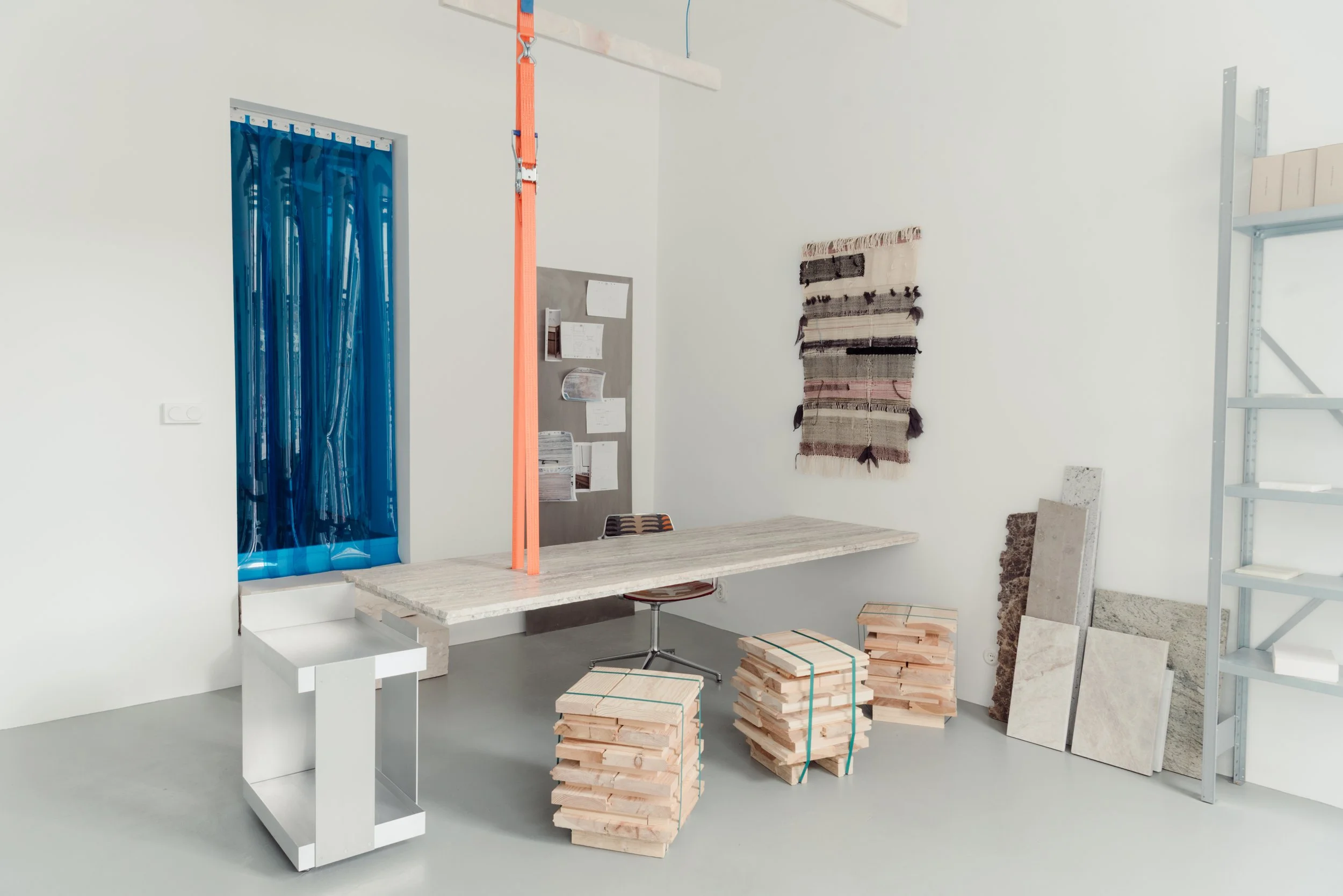 An interior room setup with a worktable, stacks of wooden planks tied with straps, a shelving unit, and various wall decorations including a woven rug. There is a blue curtain covering a window and a notice board on the wall.
