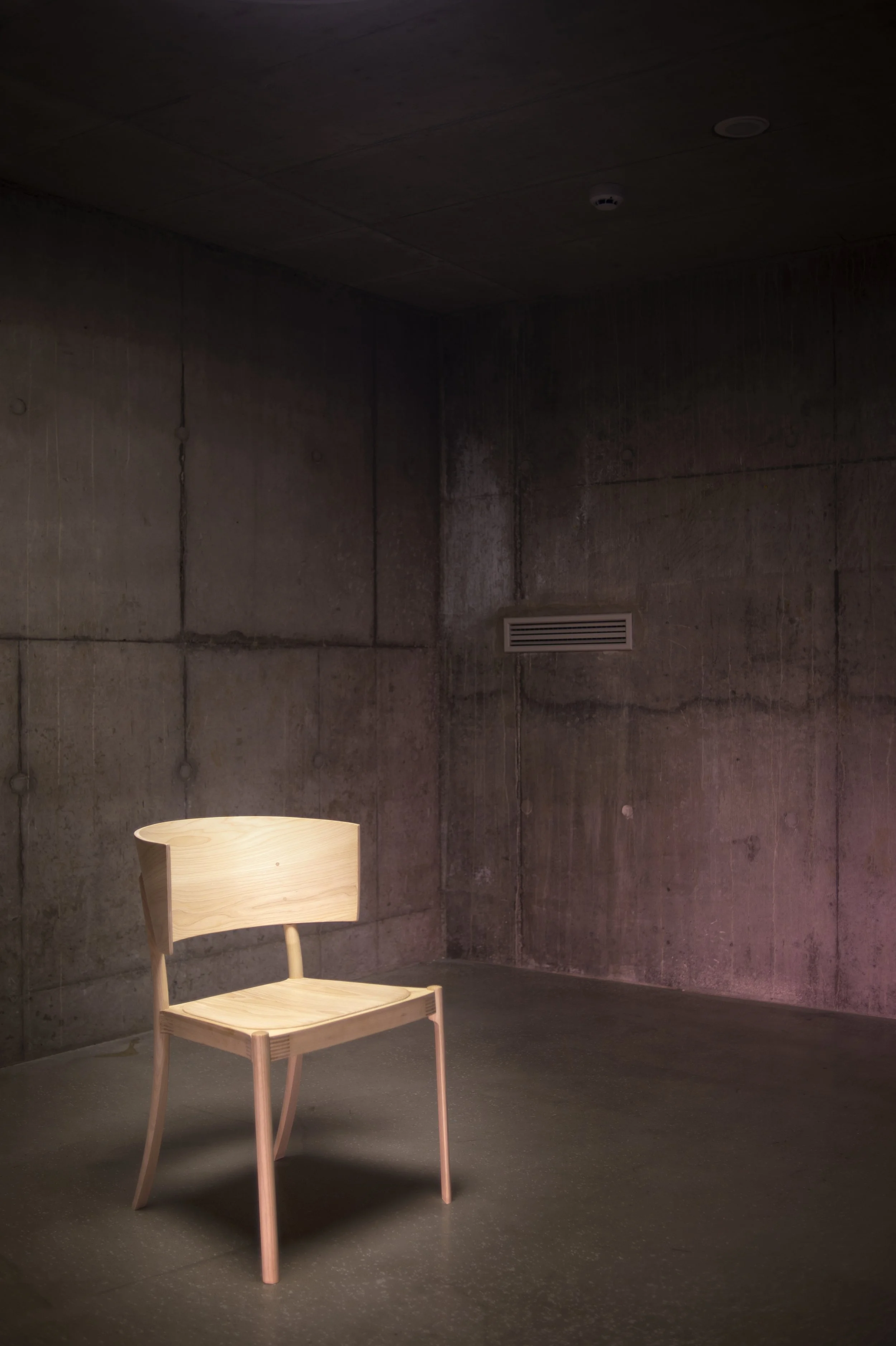A minimalistic wooden chair with a curved backrest and four slightly angled legs, placed against a dark, plain wall with visible seams and texture. A wooden floor lamp with a conical shade is partially visible in the lower left corner of the image.