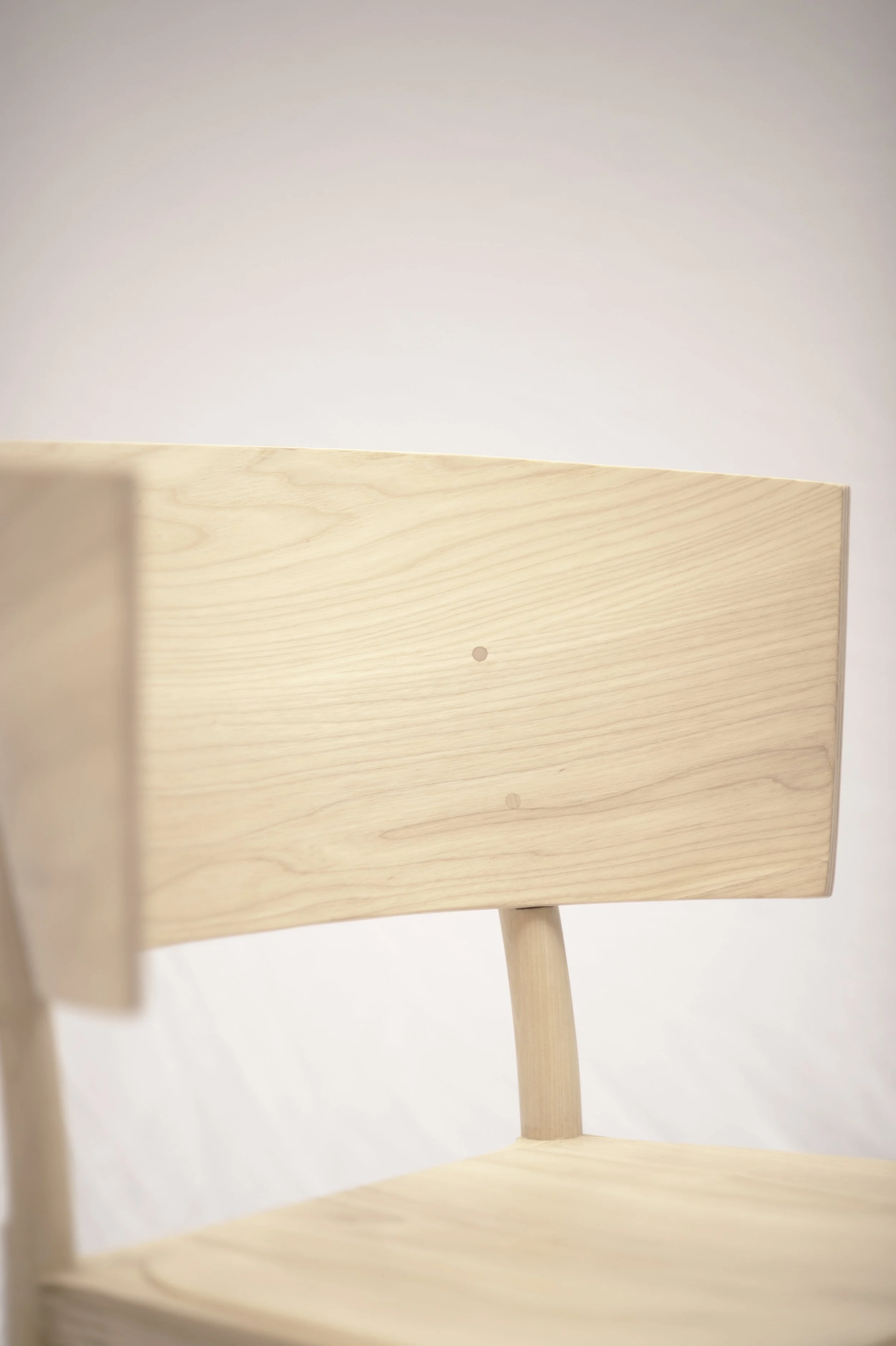 Close-up of a light wooden chair with a simple design, against a plain, light-colored background.