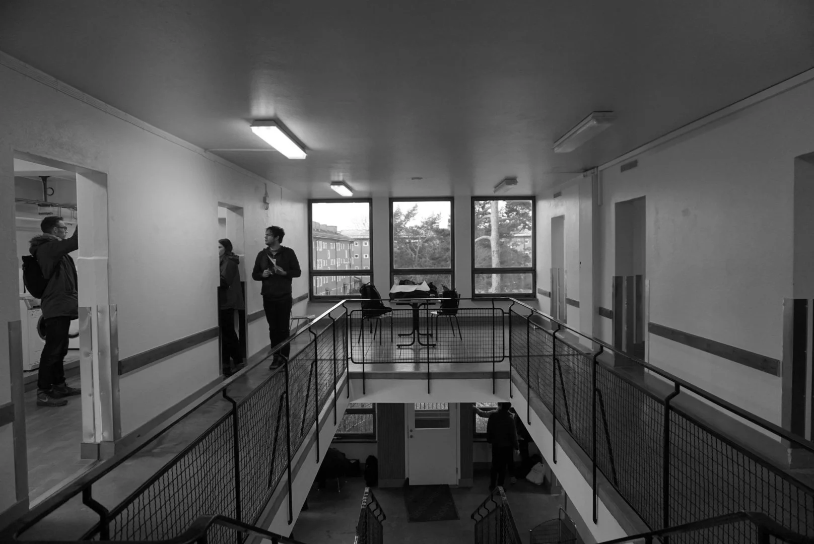 Interior shot of a multi-level building with people talking near doorways, large windows in the background, and a table with chairs on an upper level.