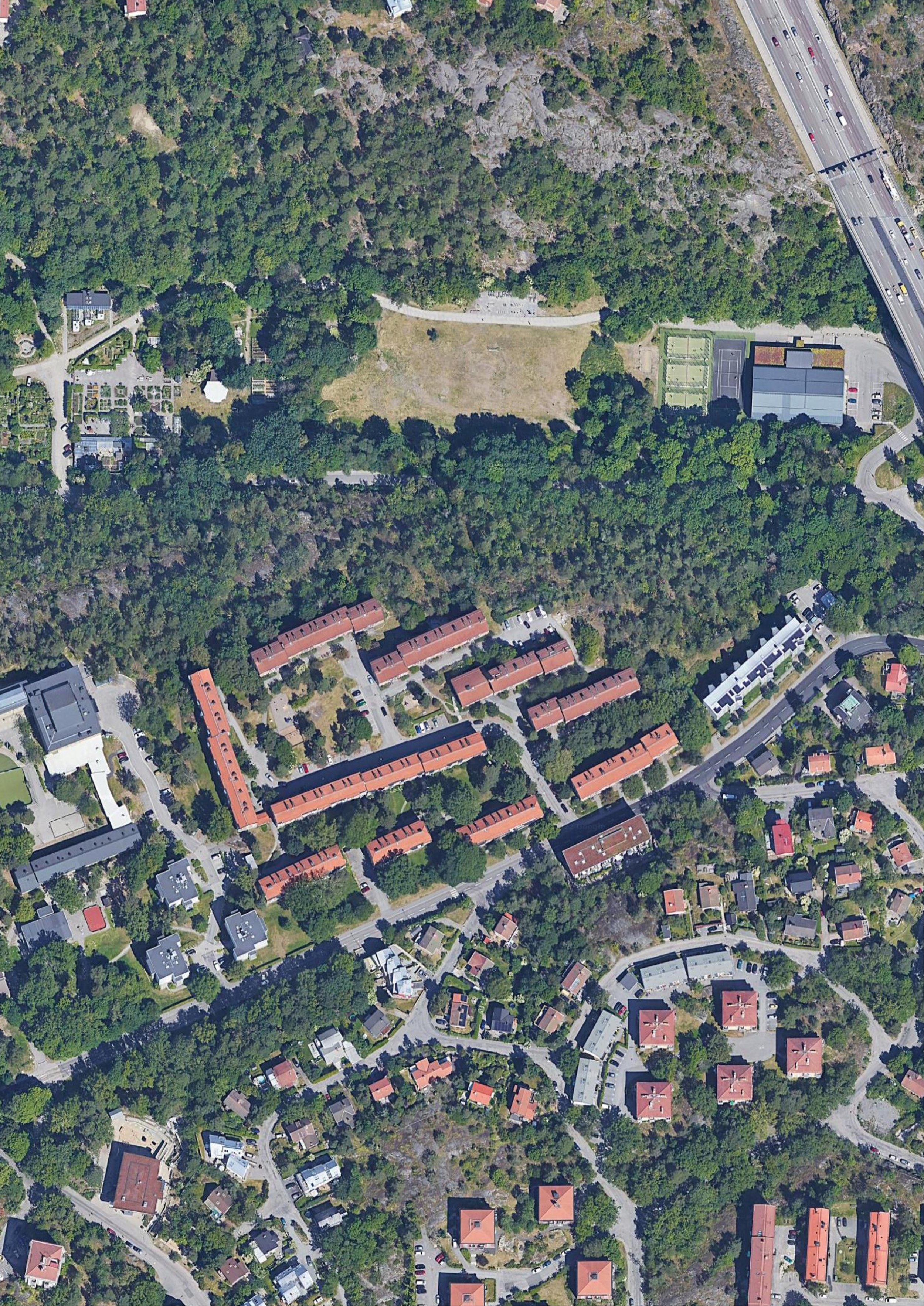 Aerial view of a cityscape with a large park in the center, surrounded by residential buildings with red roofs and a highway to the right.