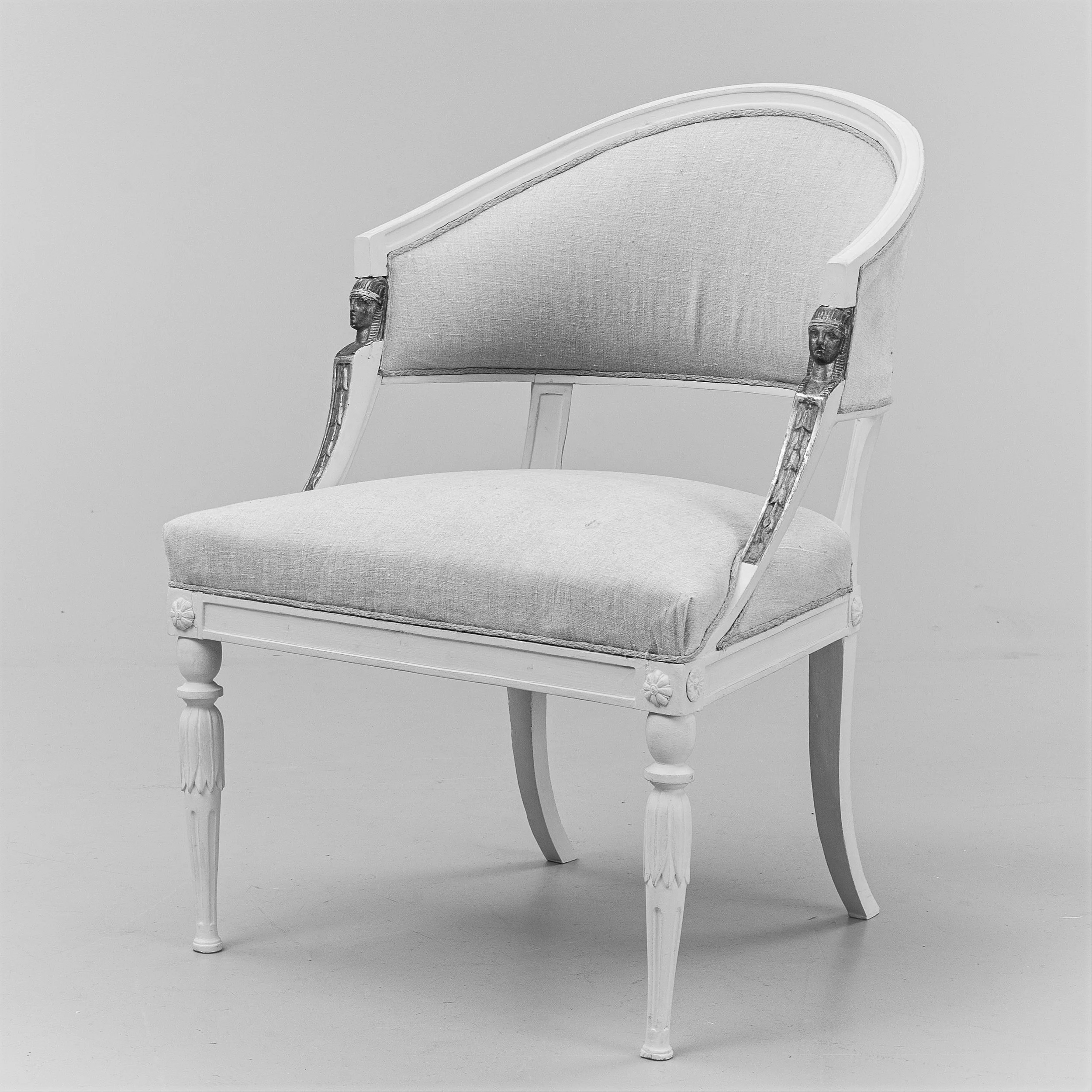 A vintage white wooden chair with carved details, including a lion's head on each armrest, a curved backrest, and turned legs, is positioned against a plain wall.