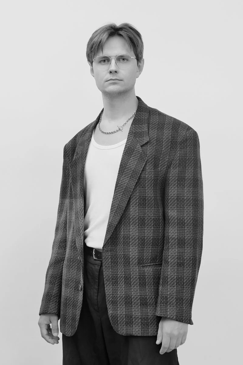 A young man with glasses, wearing a plaid blazer over a white t-shirt, with necklaces, standing against a plain background in a black-and-white photo.