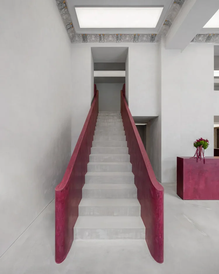 Interior staircase with white steps and red side panels, leading to an upper level in a modern, minimalist space with white walls and ceiling, and a flower arrangement on a red pedestal.