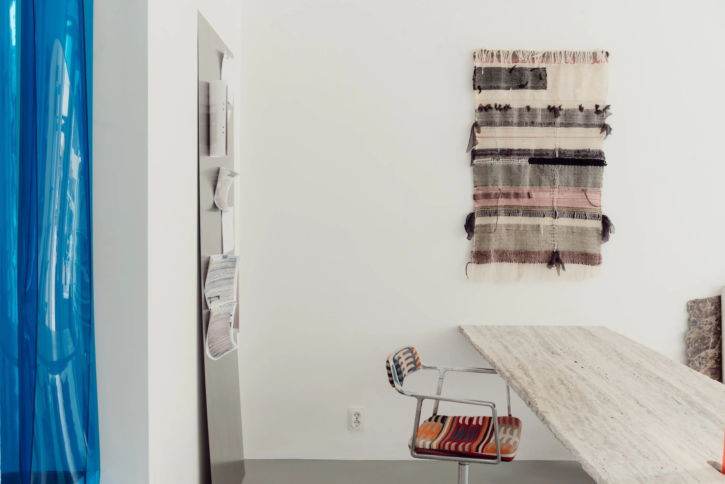 Interior of a room with a white wall, a hanging woven textile with black, cream, and pink stripes, a marble countertop, a white plastic chair with a colorful striped cushion, a small rack with magazines or brochures, and a blue curtain on the left si