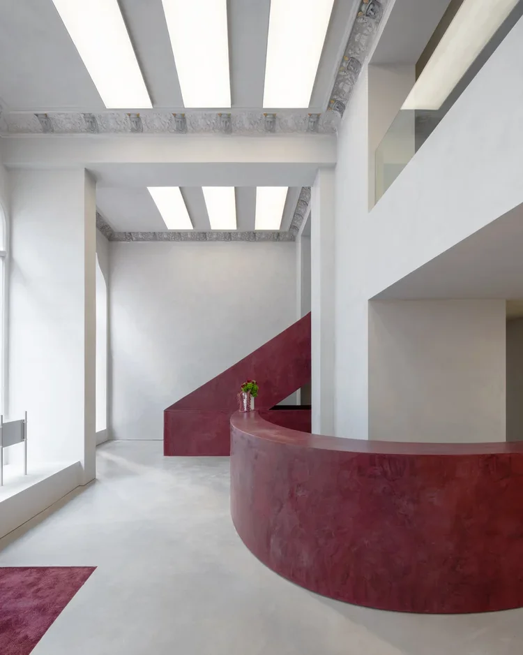 Modern interior space featuring a curved red wall and staircase with a small potted plant on top, white walls, high ceilings, and large windows allowing natural light.