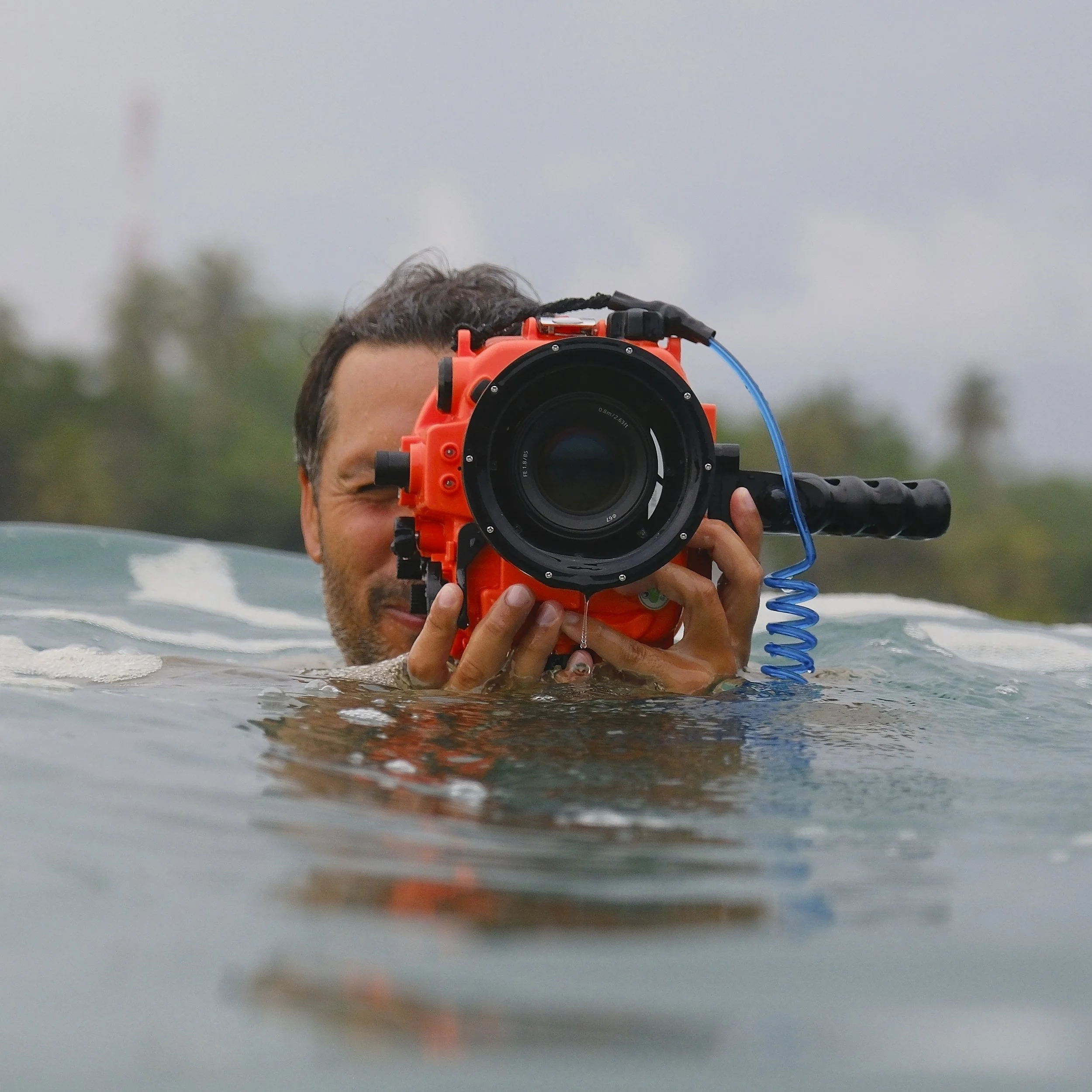 A man partially submerged in water, holding a large underwater camera with an orange housing and recording equipment.
