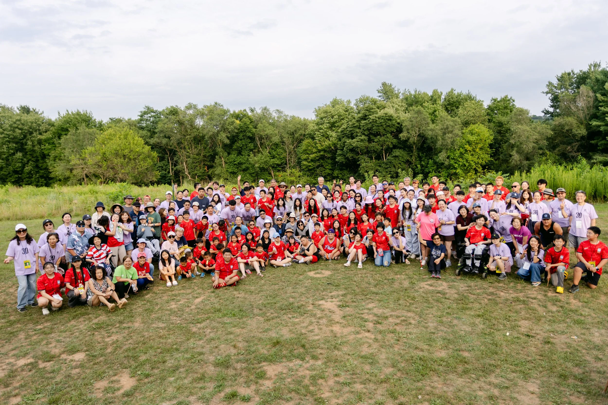 Large group of people outdoors on a grassy field, with a backdrop of green trees and a partly cloudy sky, participating in a group photo.