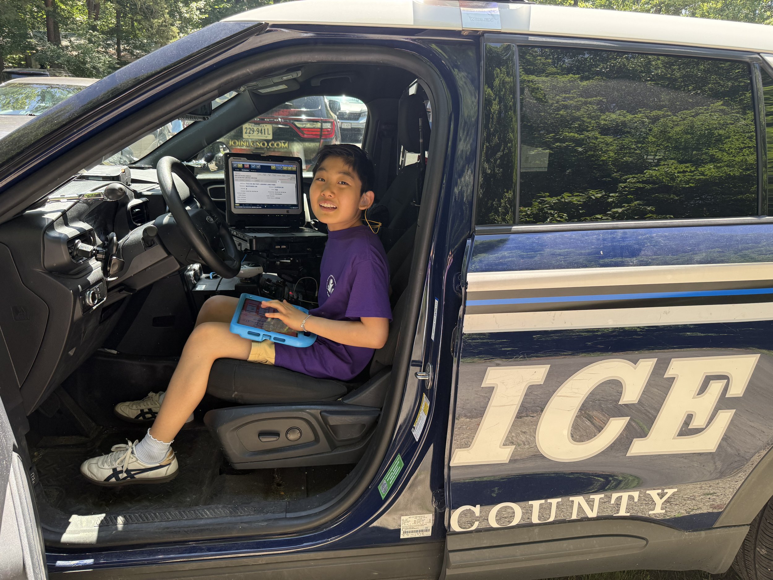 A young boy sitting inside a sheriff's vehicle, holding a tablet, with a computer monitor in front of him, smiling and looking at the camera. The vehicle has county markings on the side.