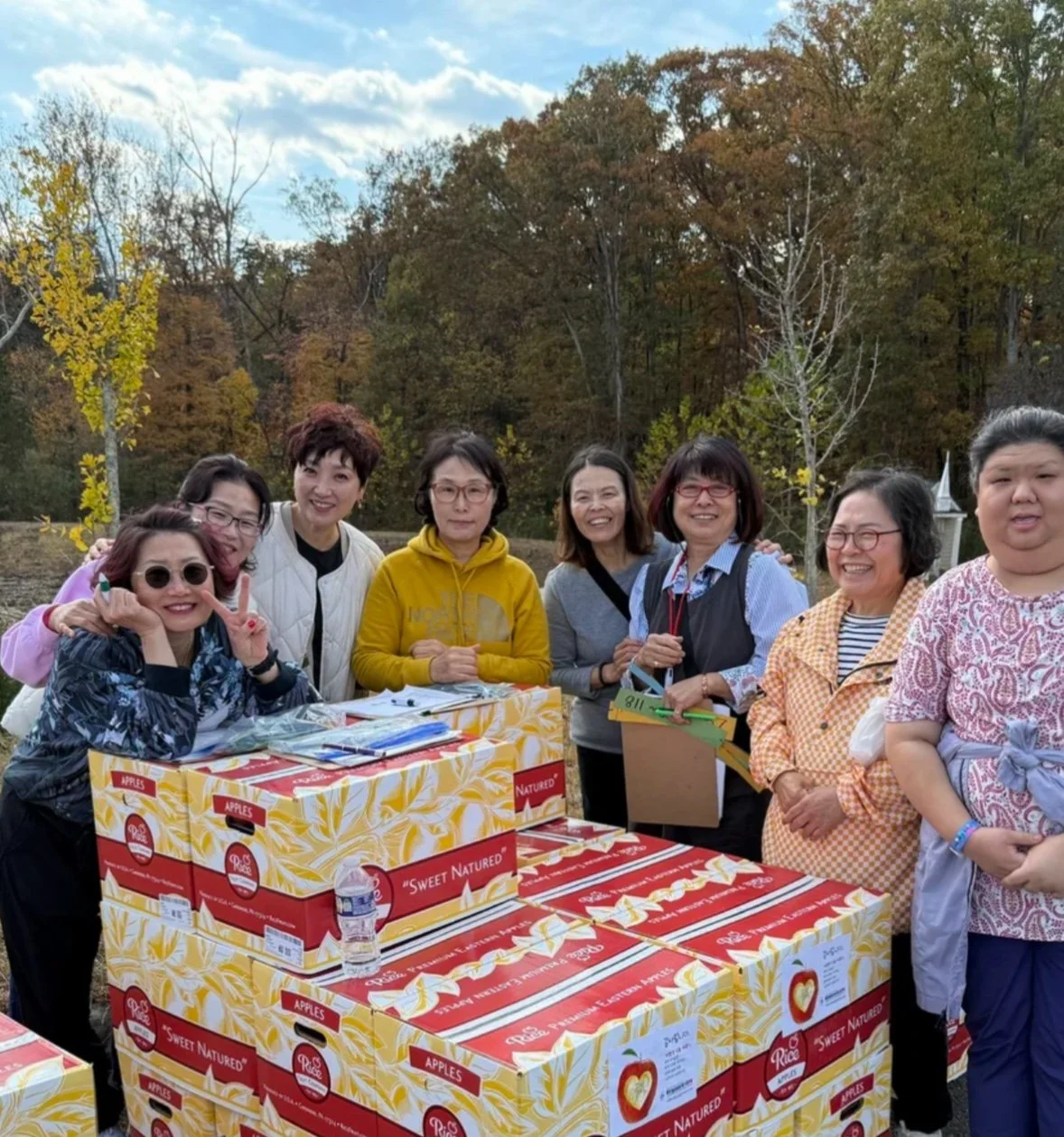 Group of women standing outdoors behind a table stacked with boxes of apples, surrounded by trees with autumn leaves.
