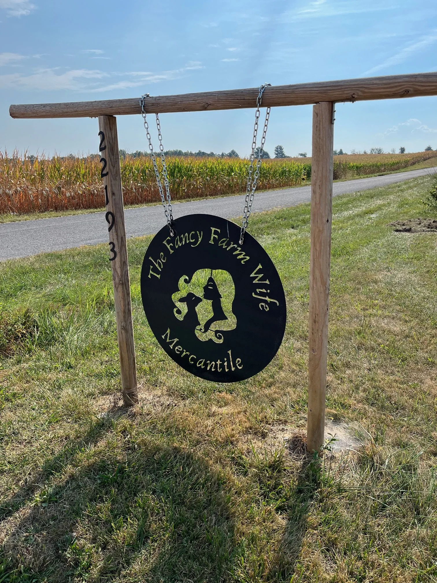 A wooden frame holding a black circular sign that reads "The Fancy Farm Wife Mercantile" with silhouettes of a woman and a dog, set against a yellow cornfield and blue sky.