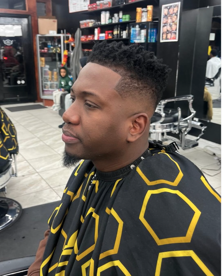Man with a fresh fade haircut sitting in a barbershop, wearing a black cape with yellow geometric patterns, with shelves of hair products and a mirror in the background.