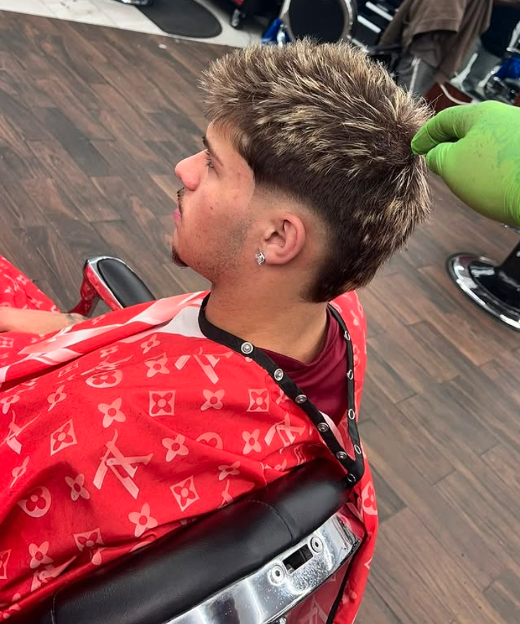 A young man with a fresh hairstyle sitting in a barber's chair at a salon, with stylists and salon equipment visible in the background, while a gloved hand holds his hair.
