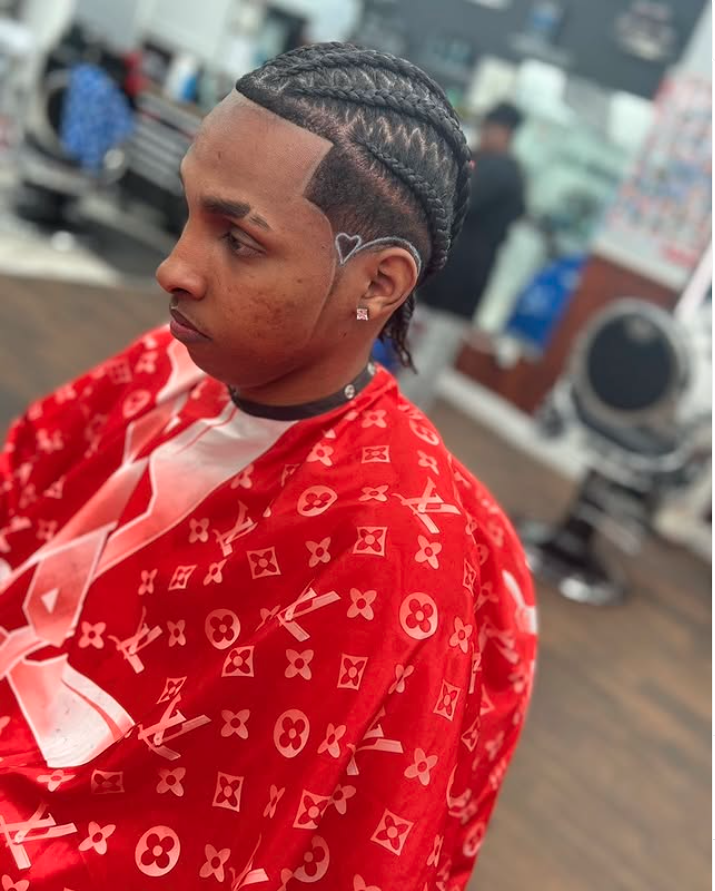 Young man with braided hairstyle, face tattoo, and diamond-shaped face tattoo, wearing a red Louis Vuitton cape, sitting in a barber shop.