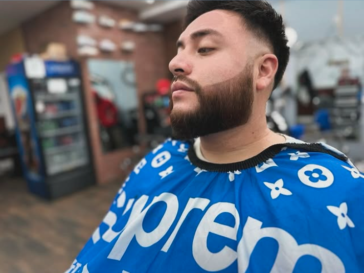 Man with a trimmed beard and short black hair wearing a blue protective cape at a barbershop or salon.