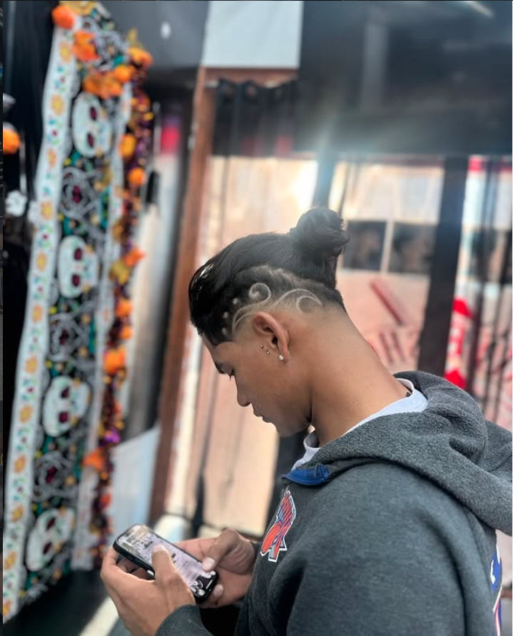 A young person with a unique hairstyle featuring shaved designs and a top bun, wearing a gray hoodie, is looking at their phone inside a store decorated with Halloween-themed items.
