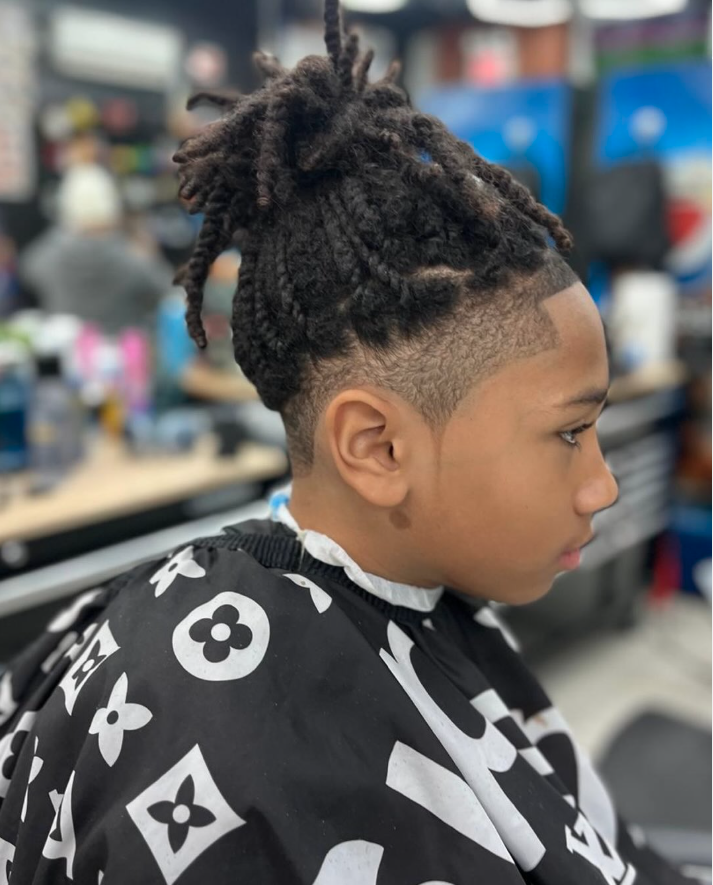 A young boy with a fresh fade haircut and dreadlocks styled in a high bun, sitting in a barbershop wearing a black and white cape with a floral pattern.