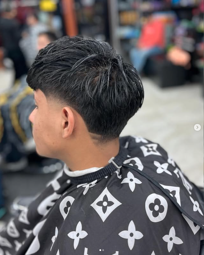 A young man with a modern haircut, featuring short sides and a longer top, sitting in a barbershop.