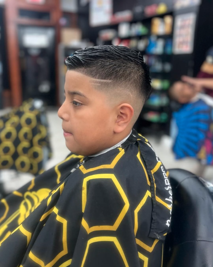 Young boy at a barbershop with a fresh haircut featuring a fade and a side design, wearing a black cape with yellow hexagonal patterns.