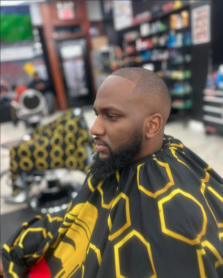 Man in a barbershop getting a fresh haircut with neatly shaped beard and a fade haircut, sitting under a cape with yellow hexagon patterns, with shelves of hair products in the background.