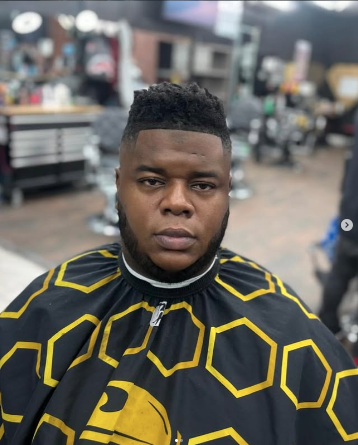A young man with a fresh haircut and beard sitting in a barbershop chair wearing a black and yellow barber cape with hexagon pattern. Barbershop interior with tools and equipment in the background.