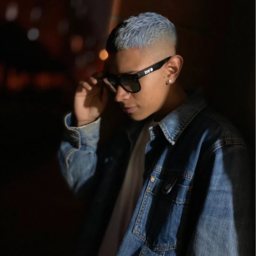 A person with short, two-tone hair (light gray at the top and darker on the sides), sunglasses, earrings, and a nose ring, wearing a denim jacket over a white shirt, tilted head slightly, with a dark background with blurred lights.