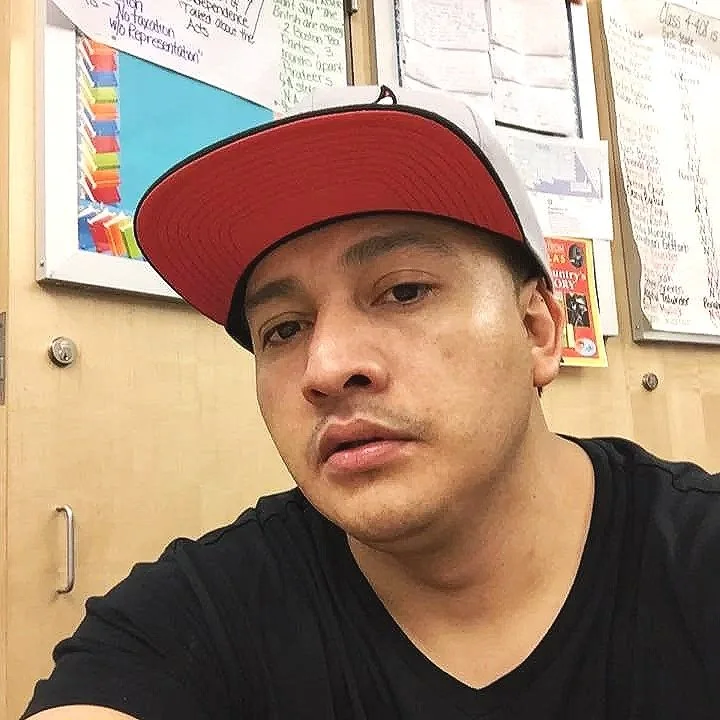 A young man wearing a black t-shirt and a red and white baseball cap, taking a selfie in what appears to be a classroom or office with bulletin boards and papers on the wall in the background.