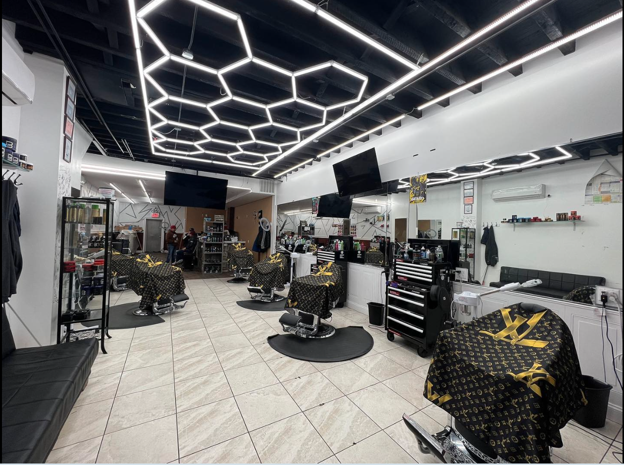 Interior of a modern barber shop with multiple barber chairs covered with black and gold Louis Vuitton print cloths, large flat-screen TVs on the walls, and a sleek ceiling with geometric light fixtures.
