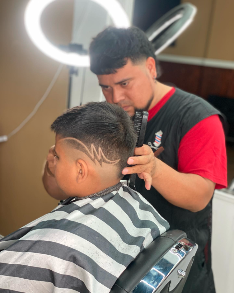 A barber is cutting a young man's hair, revealing a geometric hairstyle with a lightning bolt pattern shaved into the side, inside a barbershop.