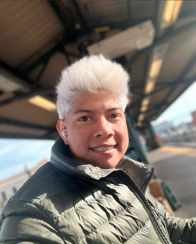 Young person with bleached white hair smiling at a train station, wearing a green quilted jacket and white earbuds.