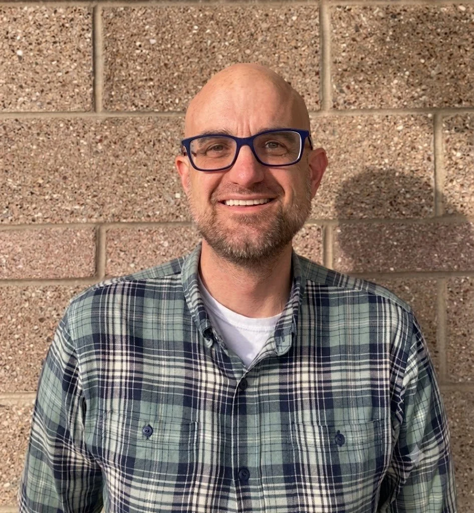 A smiling bald man with glasses and a beard, wearing a plaid shirt, standing in front of a brick wall.