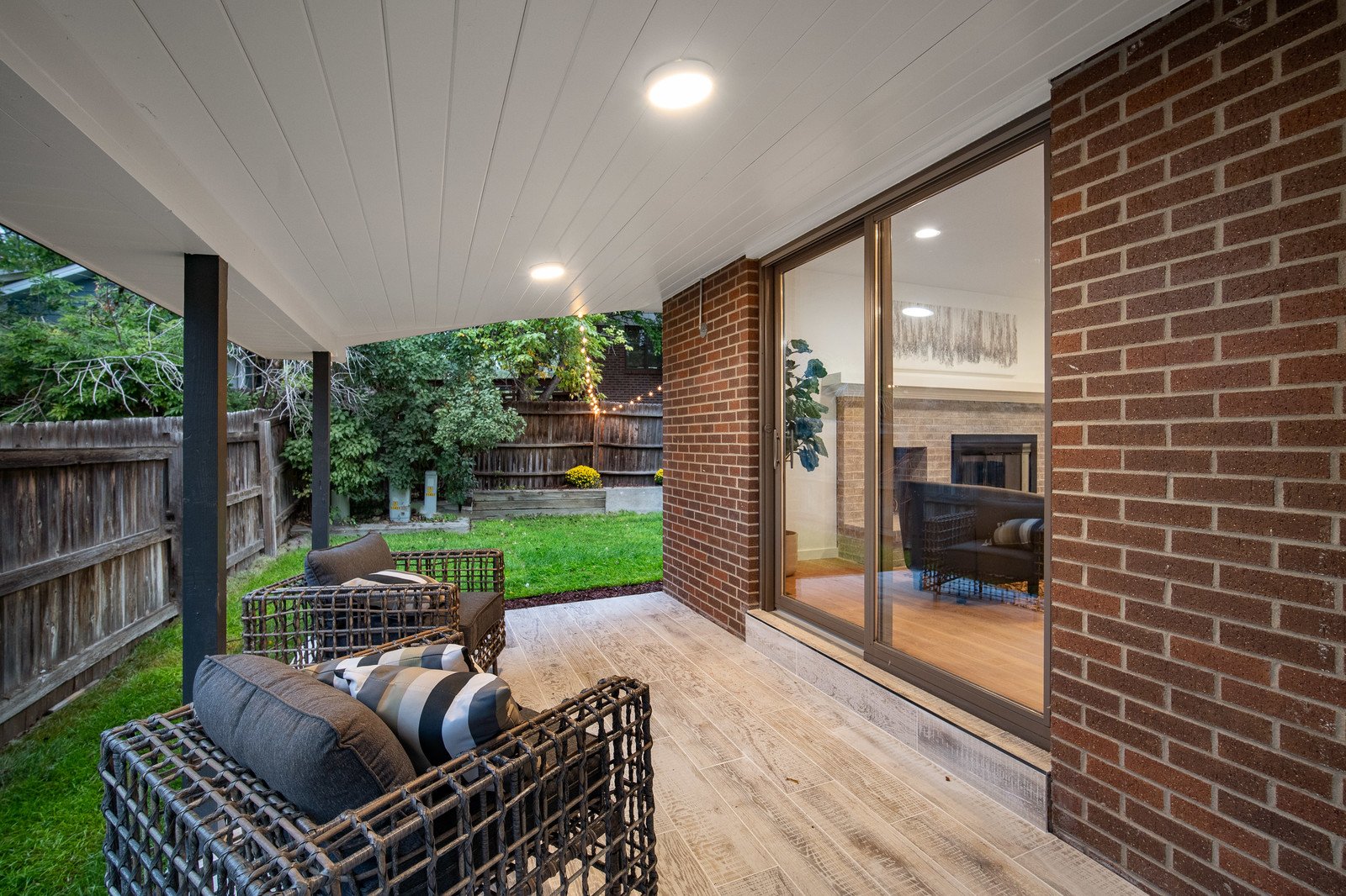 Covered backyard patio with wicker chairs, cushions, and a view of a grassy yard enclosed by a wooden fence.