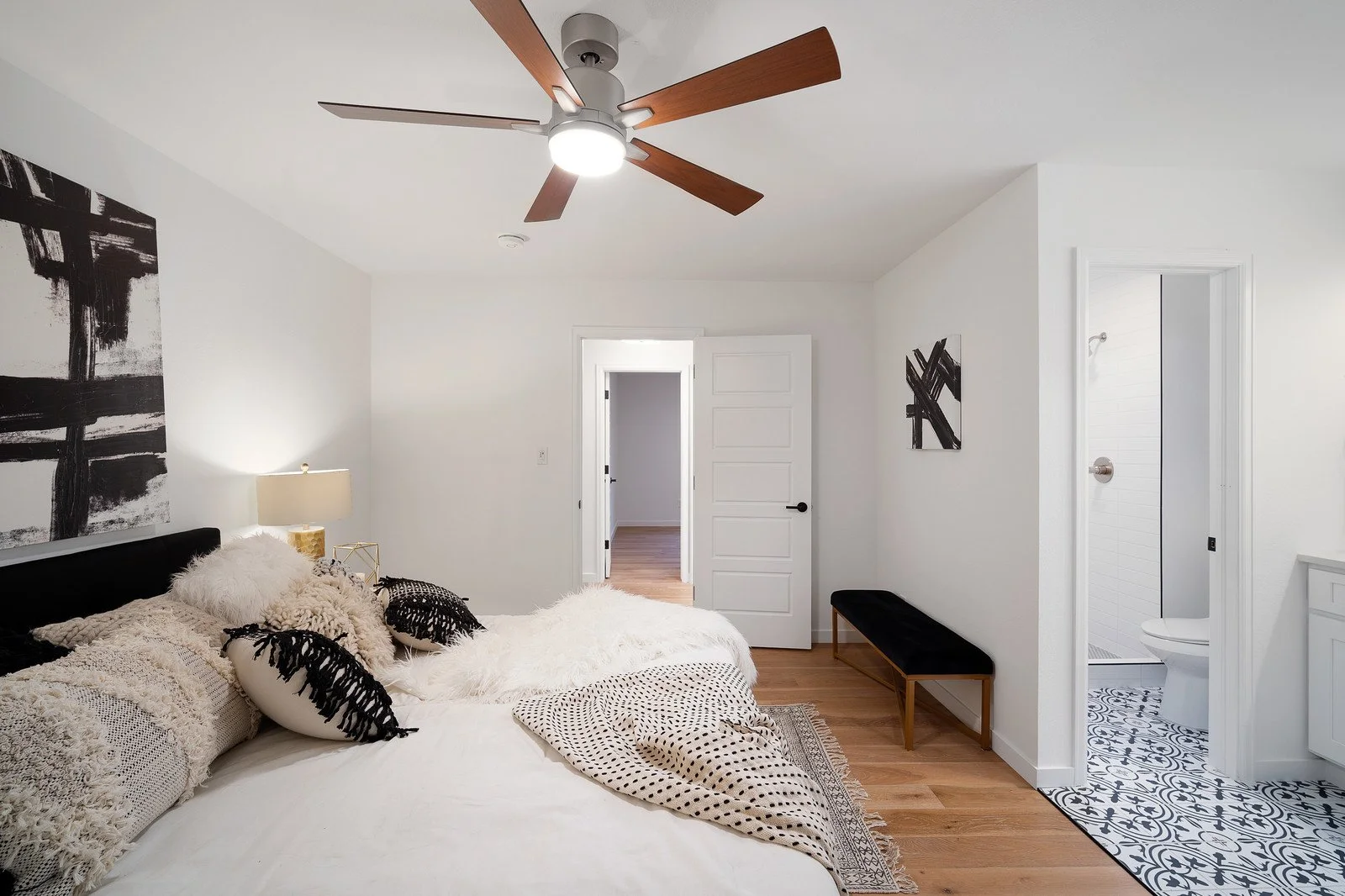 A bedroom with white walls and a ceiling fan with wooden blades. There is a bed with various textured pillows and throws, a nightstand with a lamp, a black bench, and two artworks on the walls. An open door reveals a hallway, and a small bathroom wit
